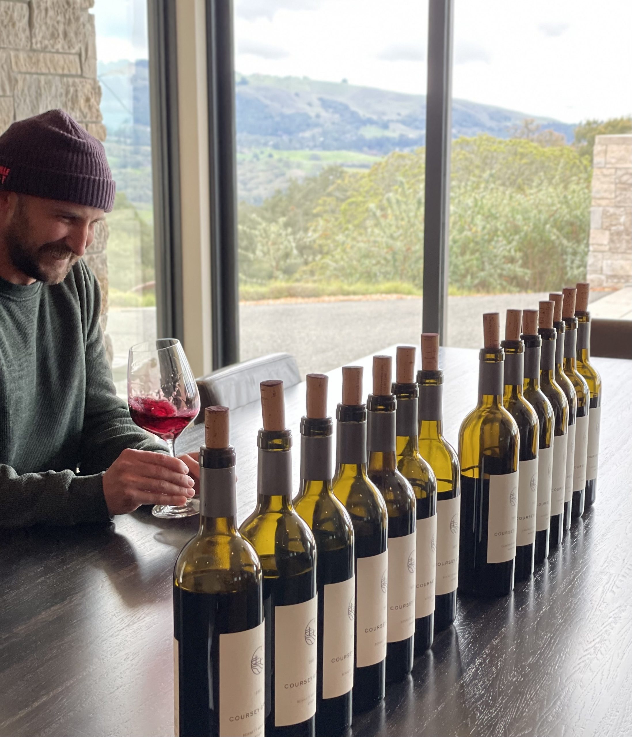 Matt swirling a glass of wine with a lineup of Coursey Graves wine bottles
